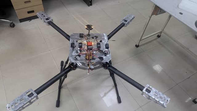 A large quadcopter standing on its landing gear on a workshop floor, with four carbon-fibre arms, machined aluminium motor mounts and an aluminium avionics deck carrying the flight electronics.