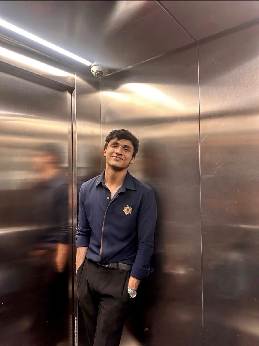 Arnav Aggarwal standing against the brushed steel wall of a lift, hands in pockets, in a navy shirt under warm overhead light.