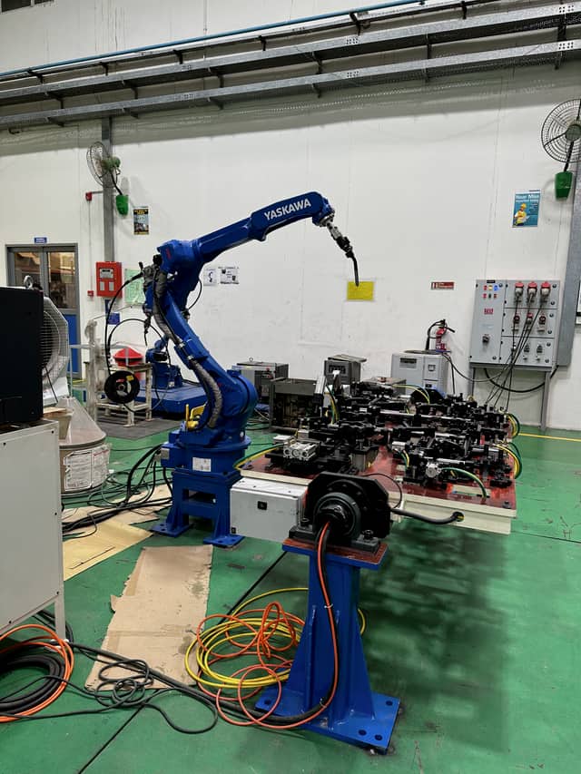 A six-axis industrial robot fitted with a MIG torch, standing over a long multi-station welding fixture on a workshop floor.