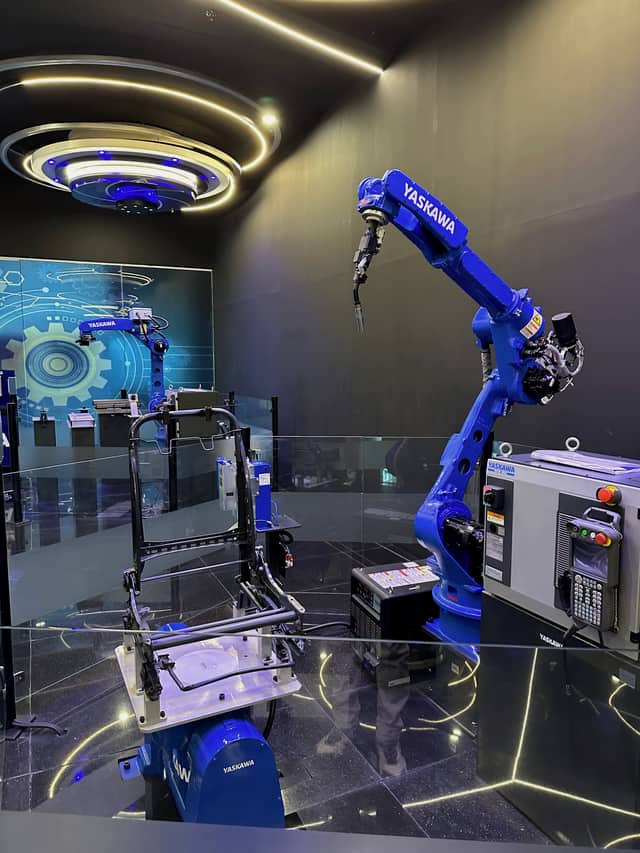 A robot with a welding torch positioned over a tubular steel frame held in a fixture, with the controller and teach pendant beside it.