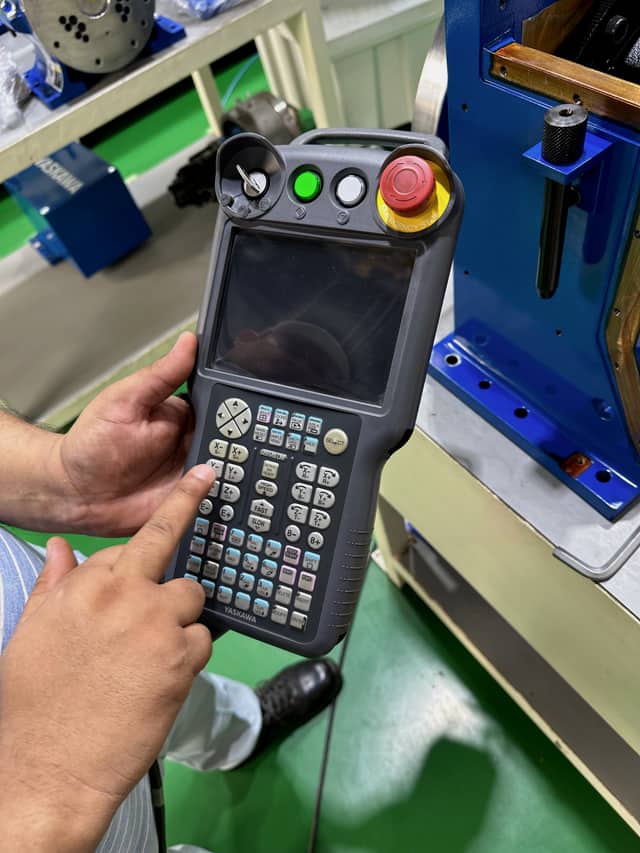 Two hands holding an industrial robot teach pendant, thumb on the jog keys, with machinery in the background.